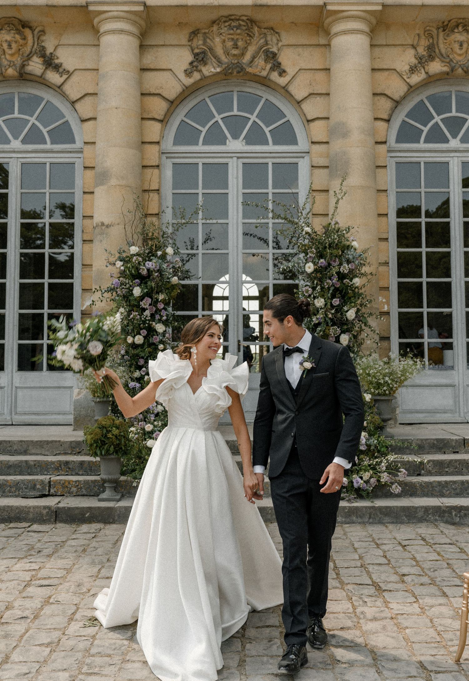 A couple walks hand in hand outside a French chateau with floral arch decorations.