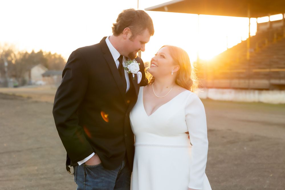A romantic couple in wedding attire share an intimate moment at sunset near a rustic outdoor venue.