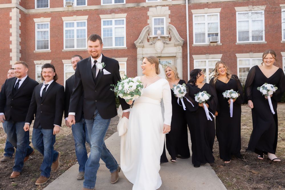 Wedding party in formal attire poses together outside brick building with bridal party in black dresses.