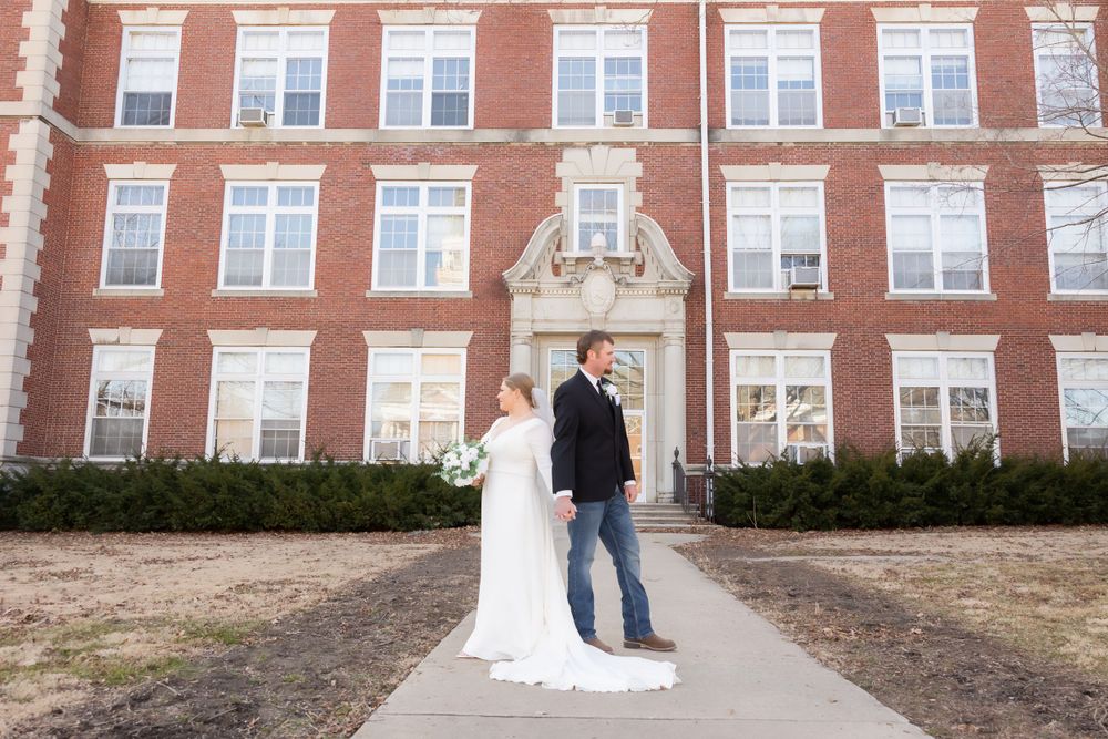 Wedding couple stands on sidewalk in front of multi-story brick building for formal portrait.