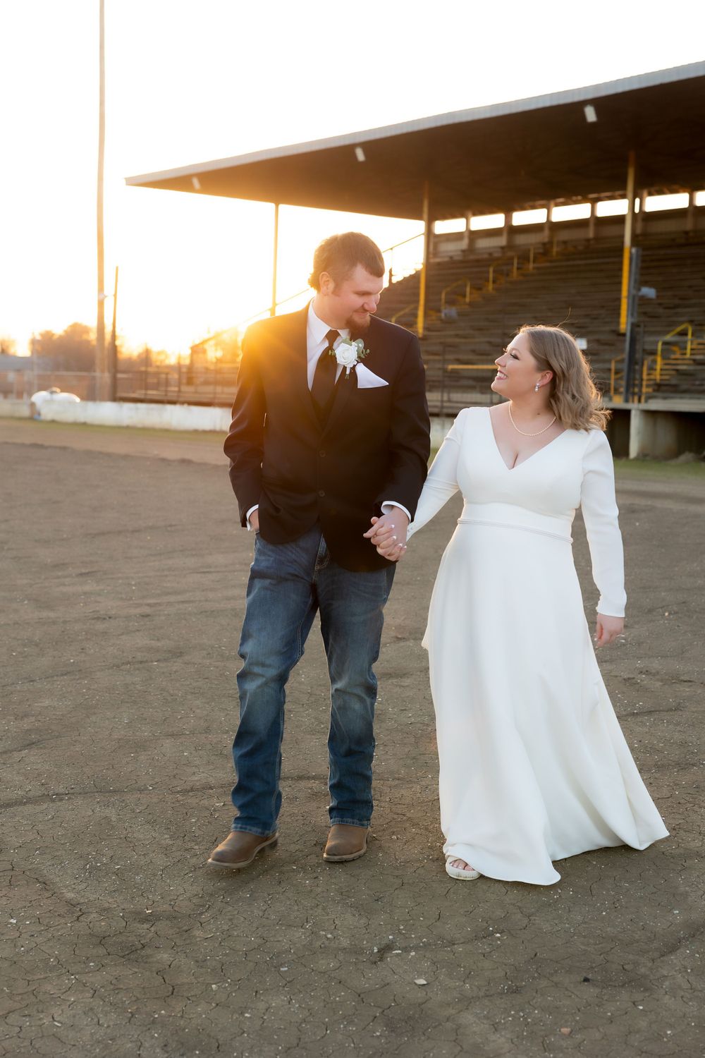 A couple walks hand in hand in wedding attire near empty stadium bleachers at sunset.