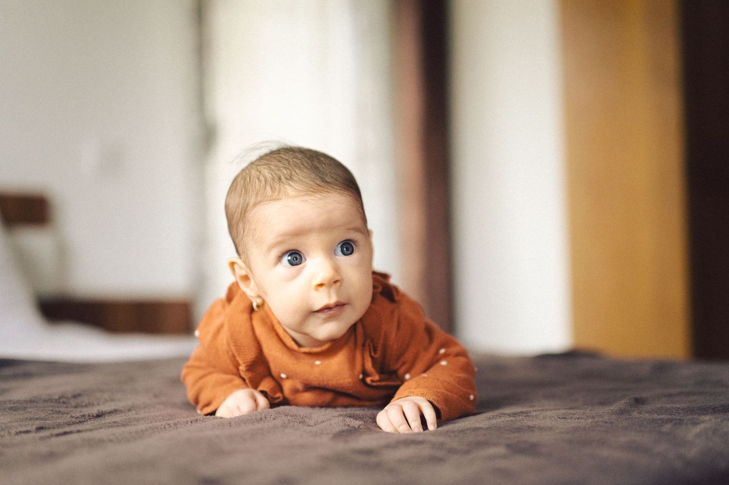 Infant in orange outfit crawling on dark bedding looking to side.