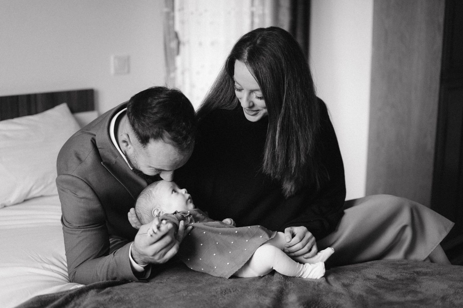 Intimate black and white photo of parents sharing a tender moment with their newborn baby on a bed.