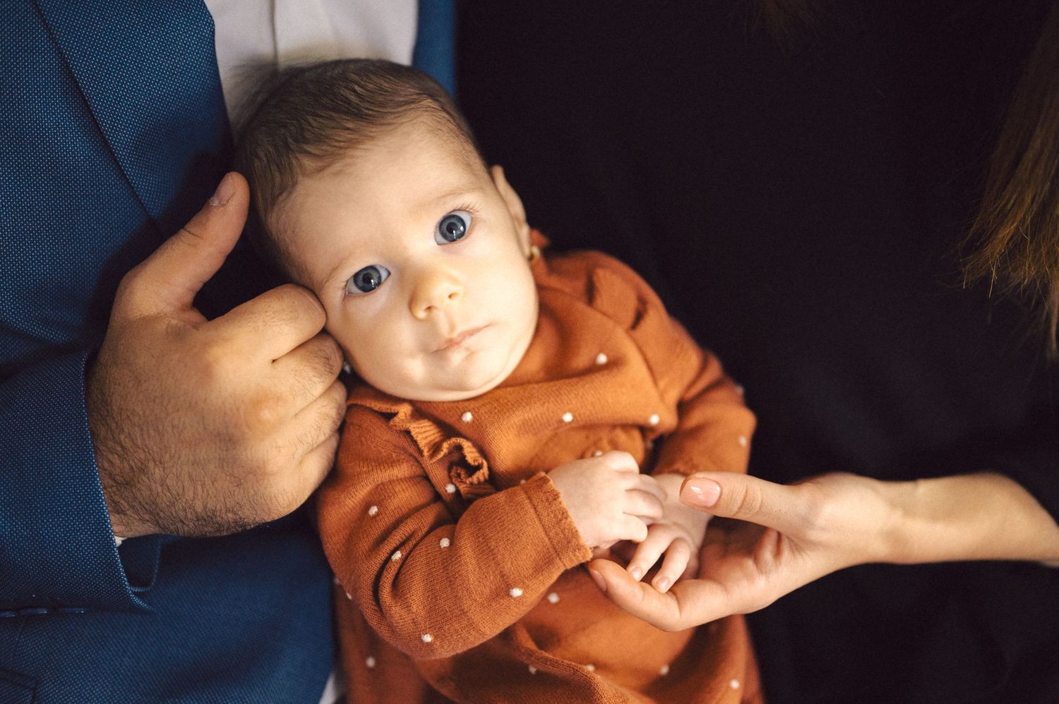 Close-up of a baby in an orange outfit with bright blue eyes being held by parents.