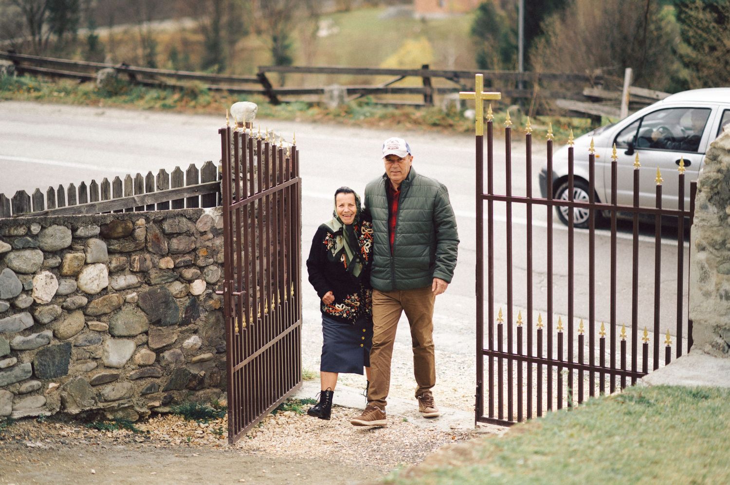Two people walking through rustic iron gates with stone wall entrance.