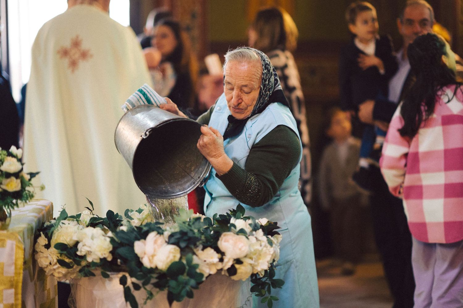 Priest pouring holy water during traditional religious ceremony surrounded by white floral arrangements.