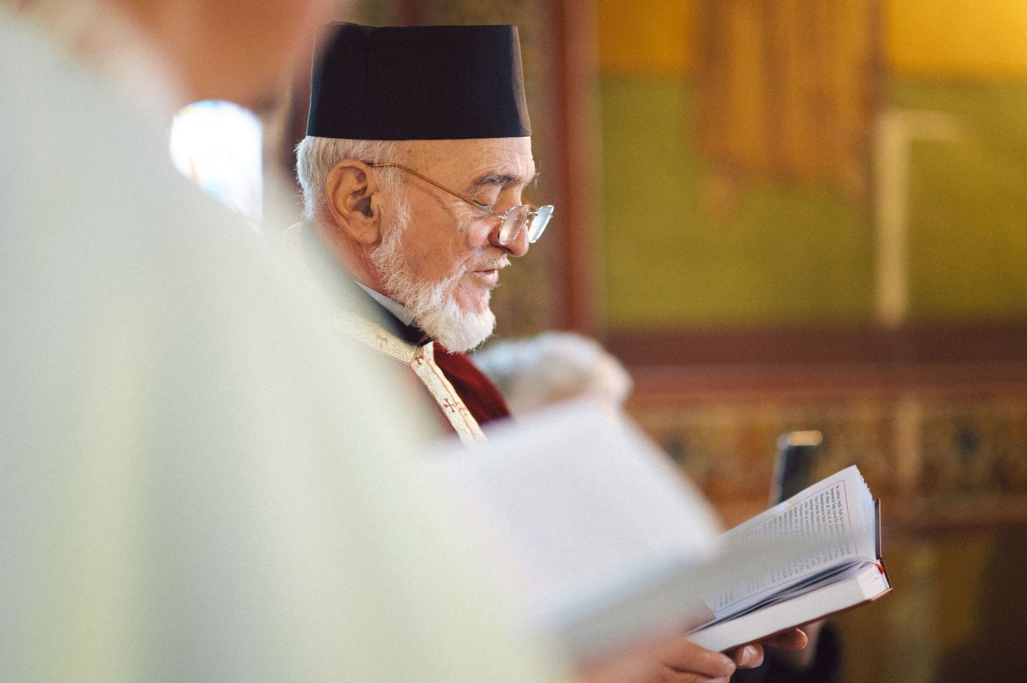 Religious leader in traditional cap reading from holy book during ceremony.