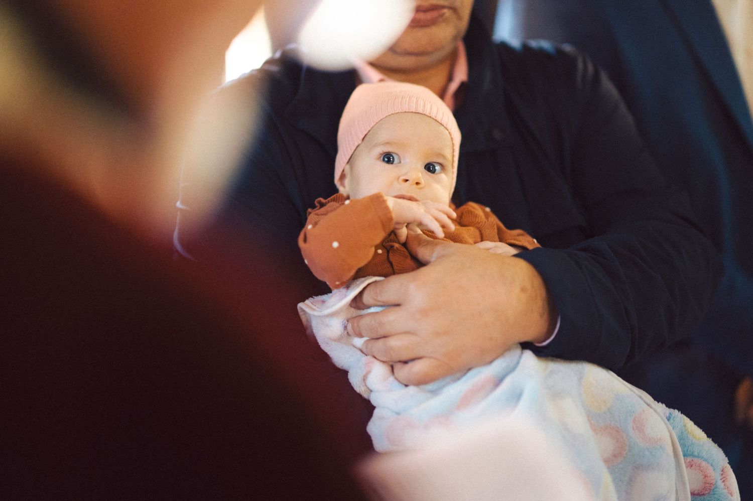 A baby in a pink hat being cradled during a baptism ceremony.