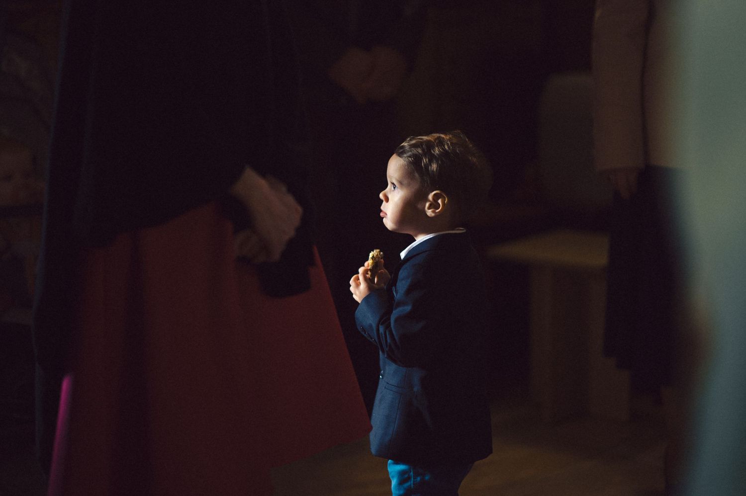 A young child in formal attire stands thoughtfully in dim lighting.
