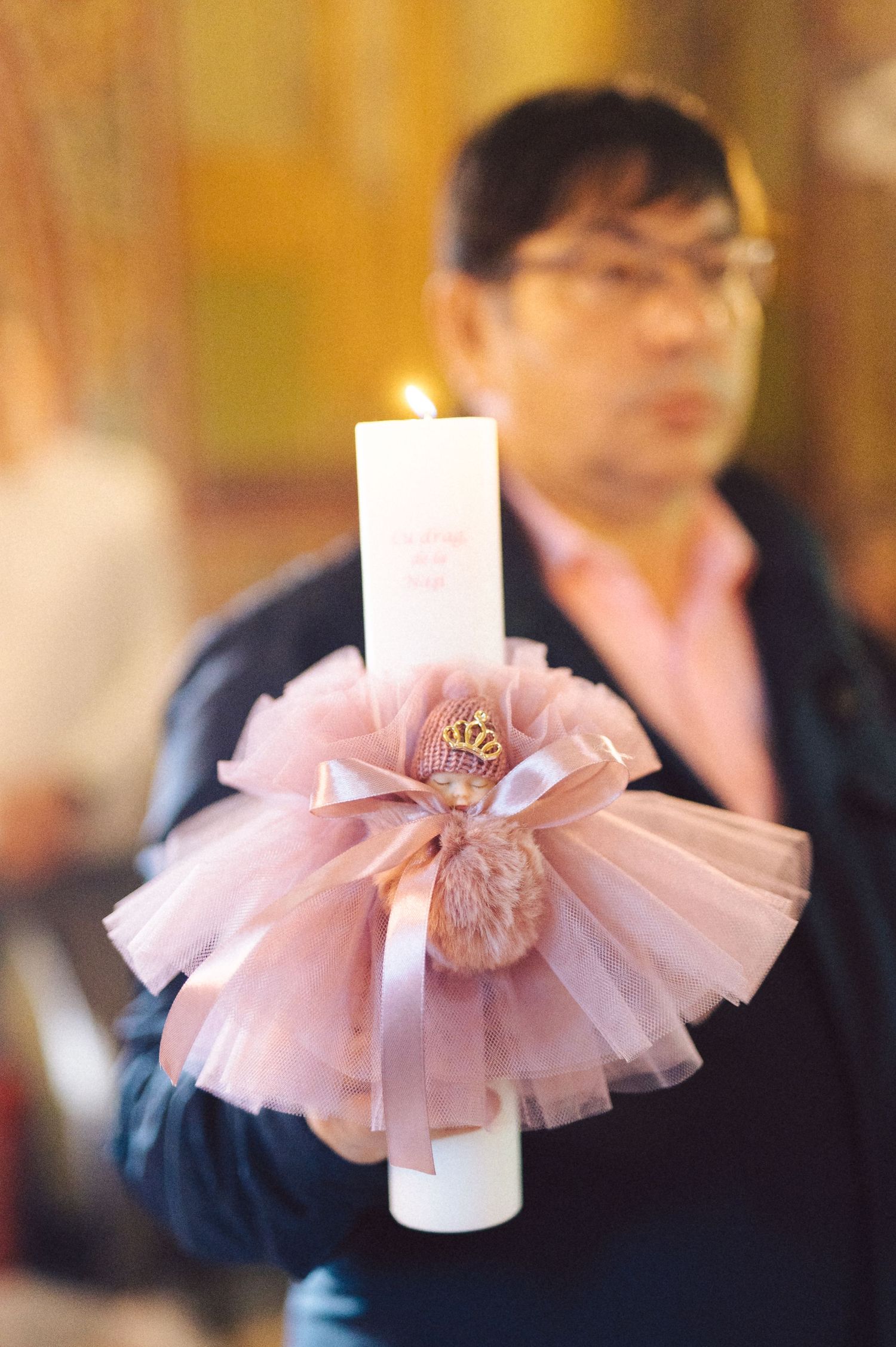 A decorative white candle adorned with a pink tulle bow and ribbon being held during a ceremony.