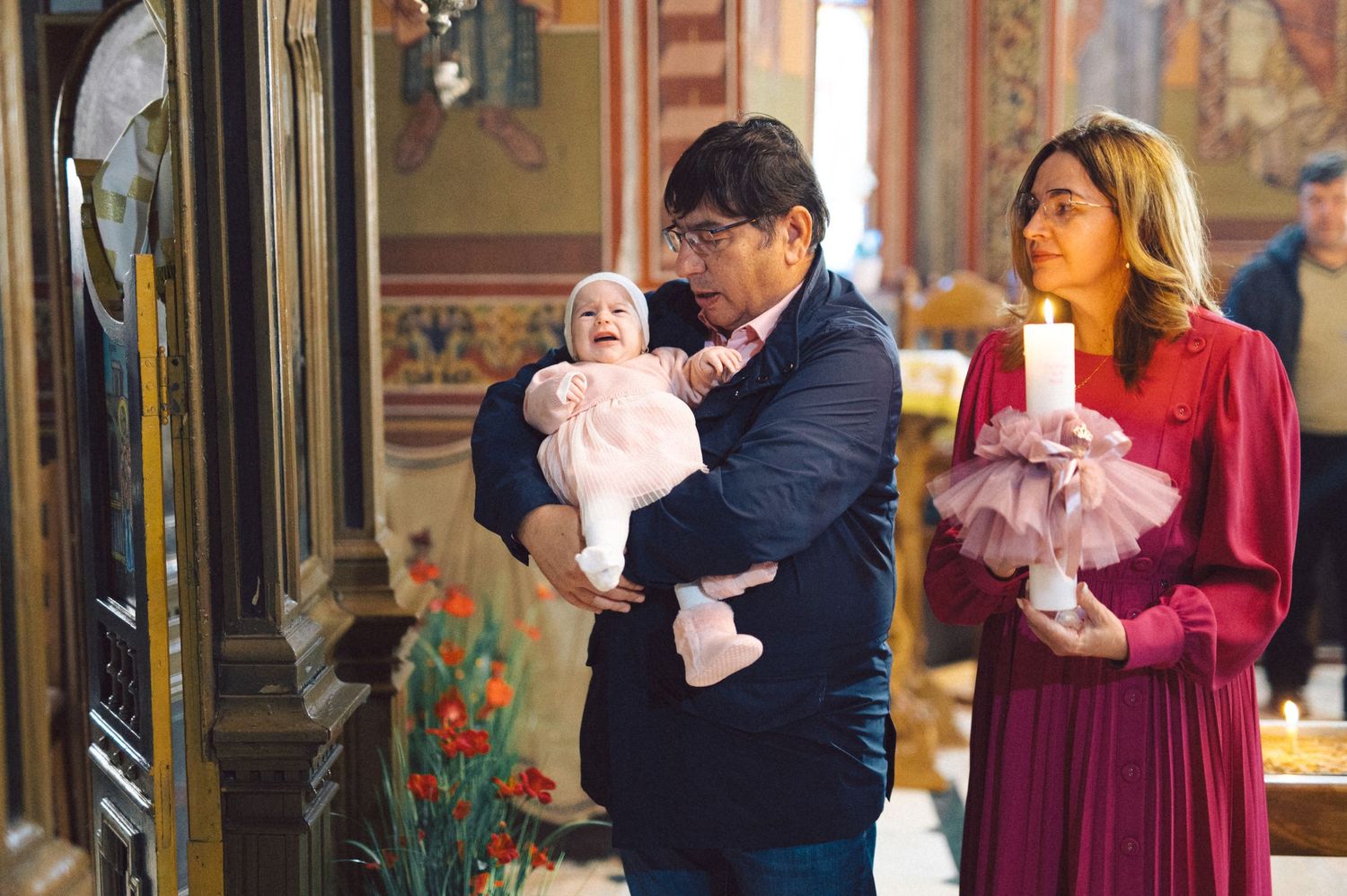 A baby in pink being held during a baptism ceremony in a decorated church.