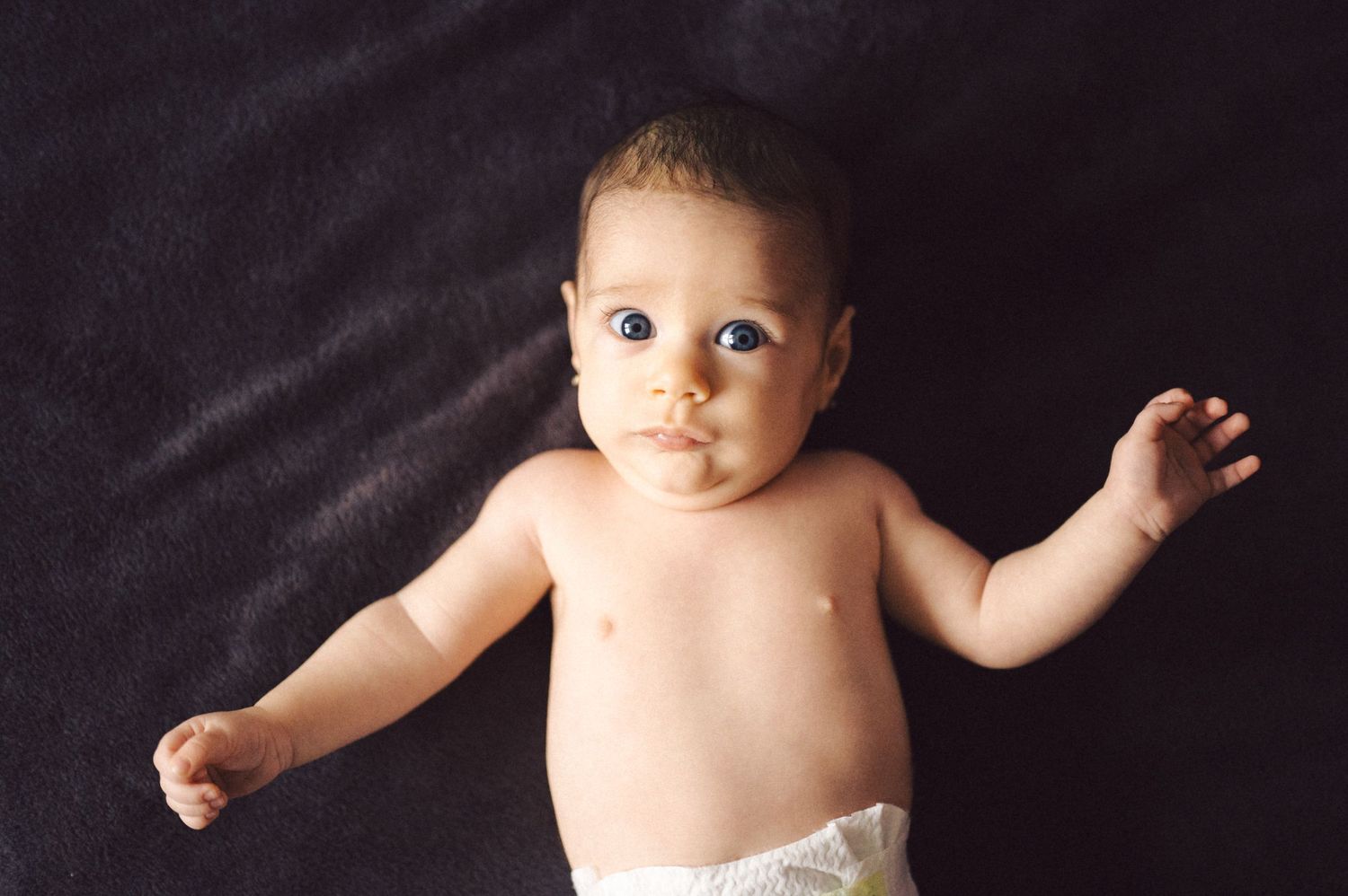 Small infant in white diaper lying on dark fabric.