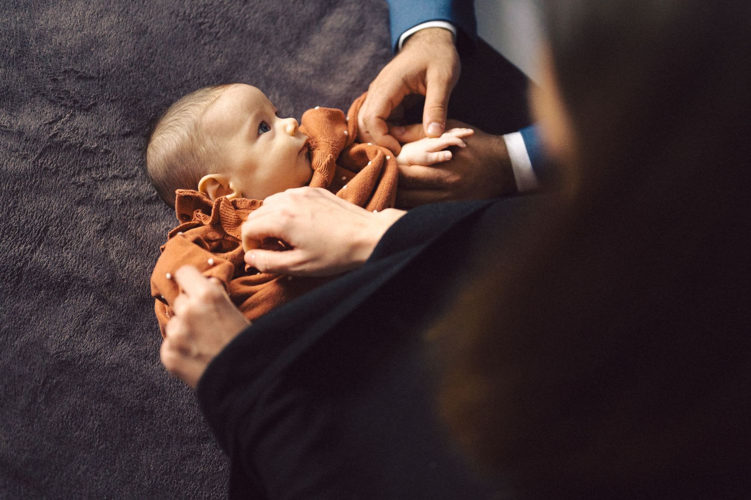 Hands reaching to dress infant in orange clothing on dark surface.