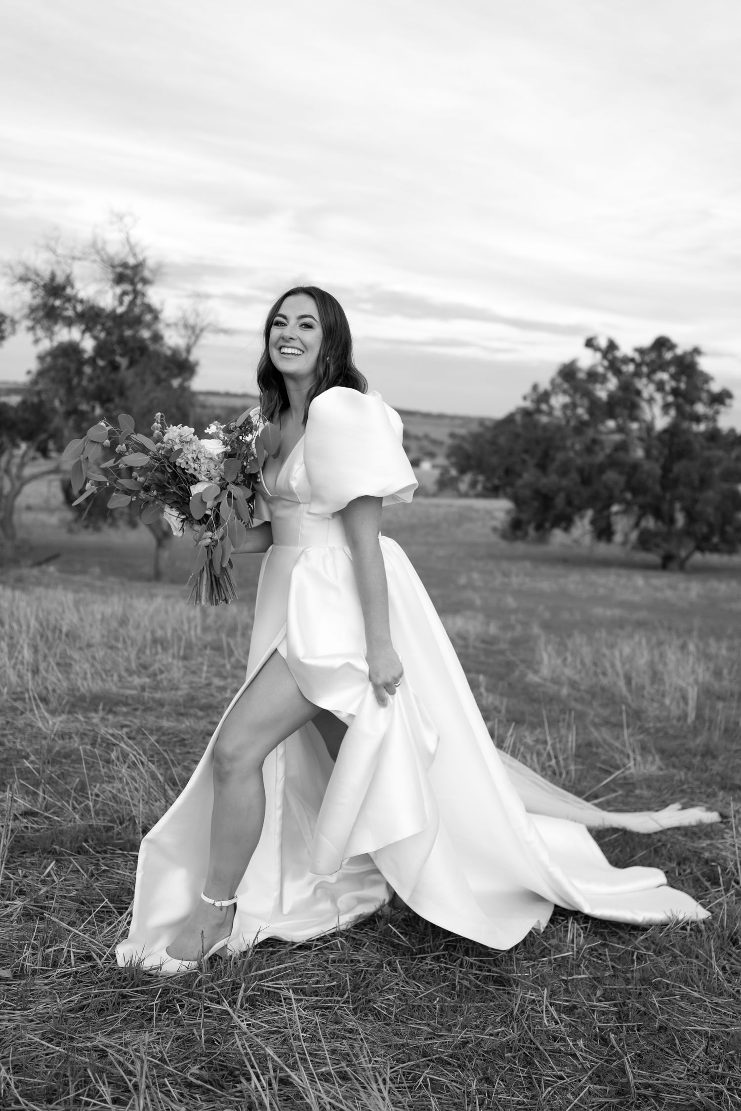 A happy figure in a flowing white wedding dress with puff sleeves holds a bouquet while standing in a scenic field at dusk.