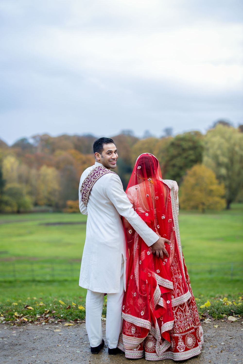 A couple in traditional Indian wedding attire poses together in a scenic autumn park.