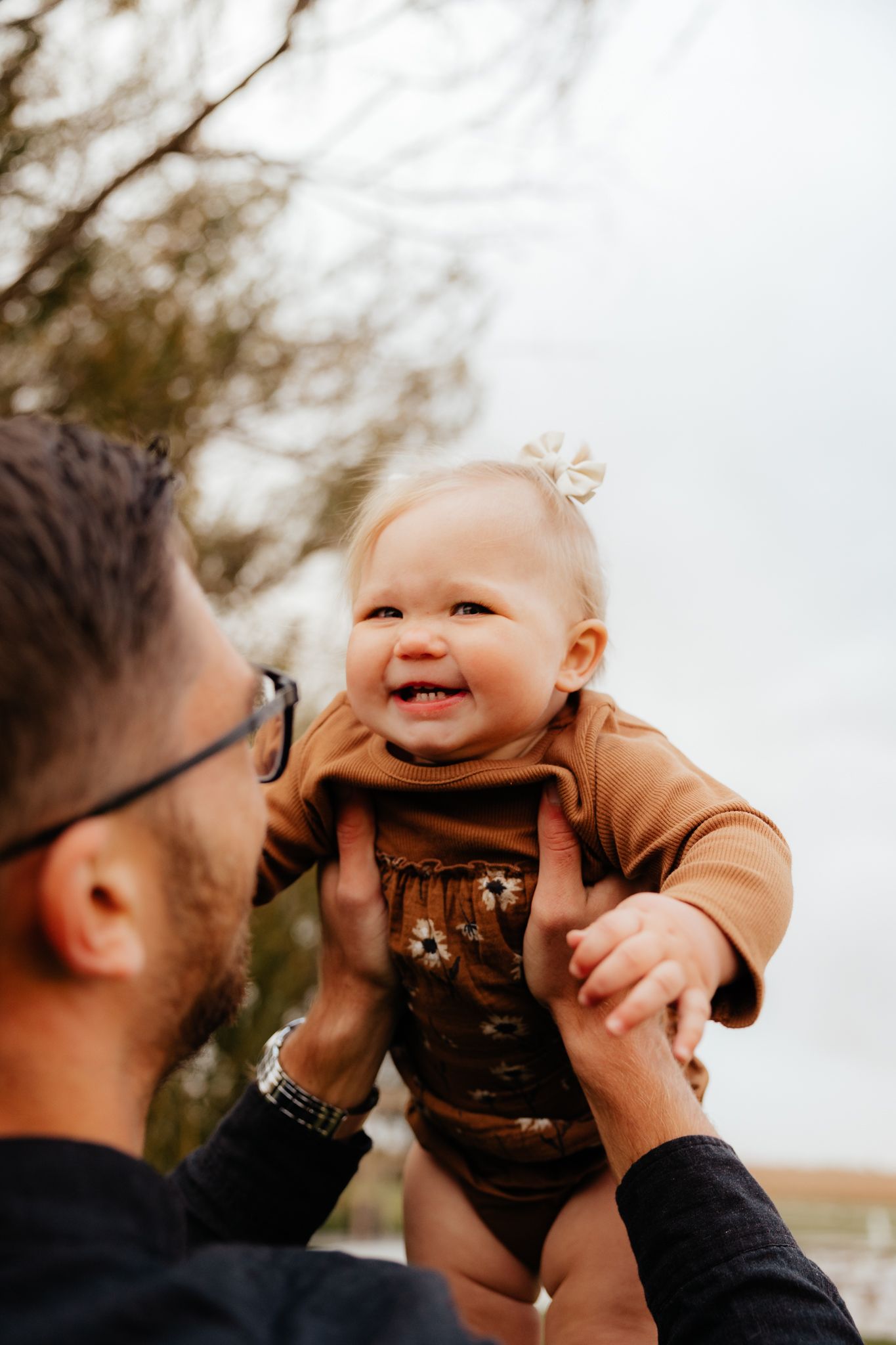 Baby in brown outfit smiles joyfully while being lifted in the air outdoors.