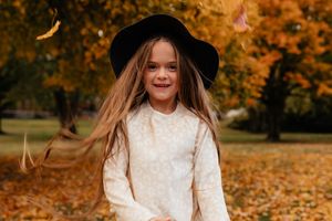 Child in white dress and black hat among fallen autumn leaves.