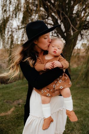 Mother in black hat shares tender moments with baby in brown outfit against natural backdrop.