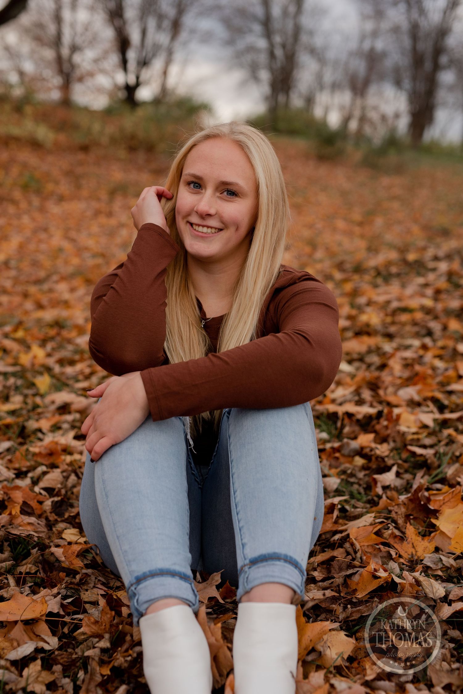 Person in brown sweater and jeans sitting among colorful autumn leaves with a warm smile.