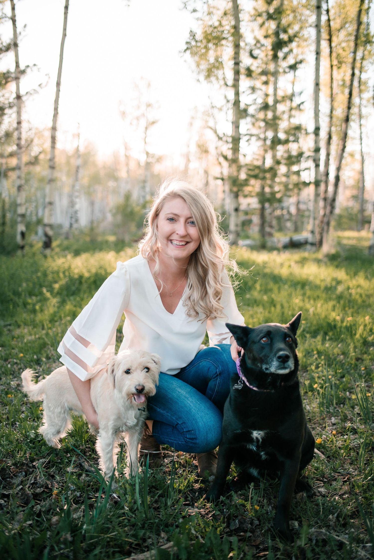 Person sitting in forest clearing with two dogs, one black and one white.