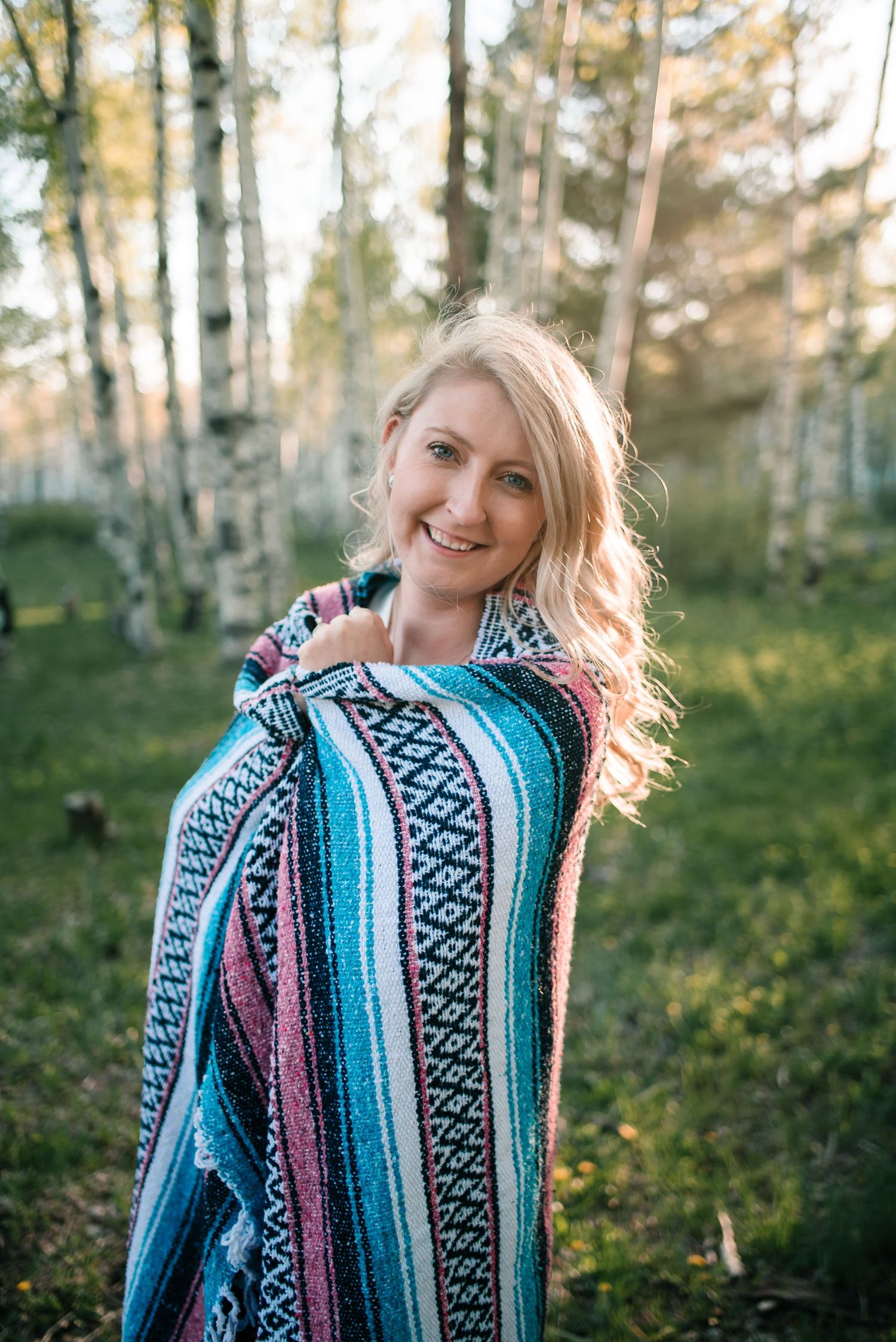 Person wearing colorful striped Mexican blanket smiling in aspen forest.