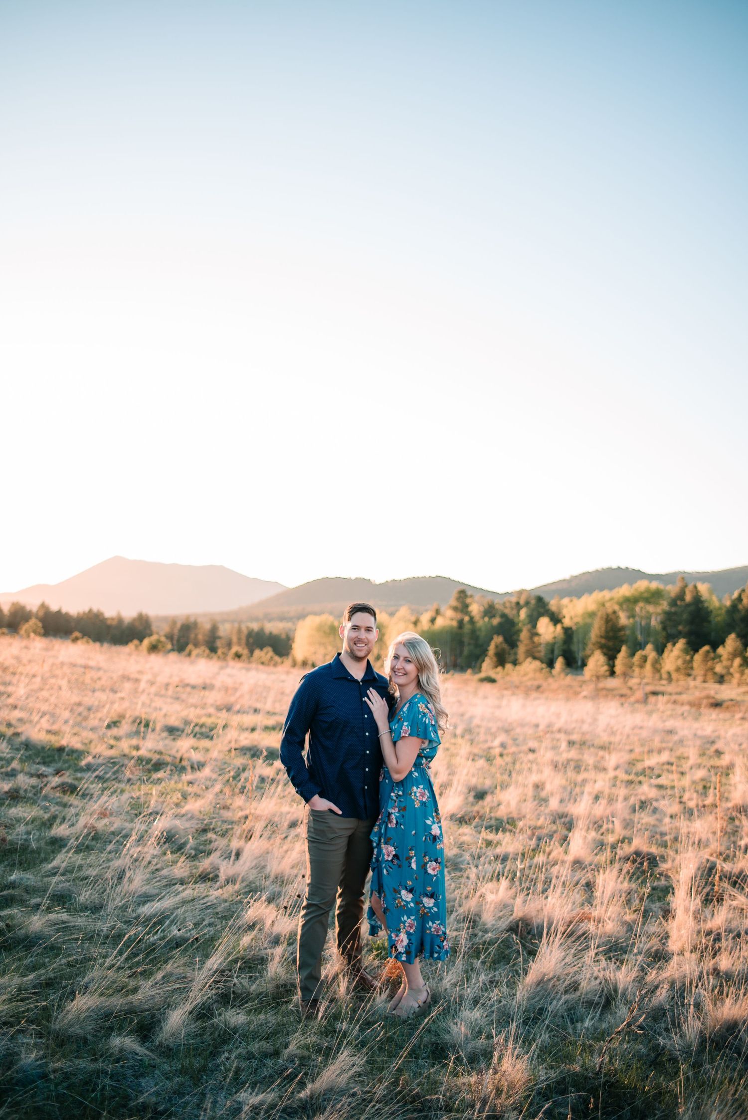 Couple standing together in field of tall grass with mountain backdrop at sunset.