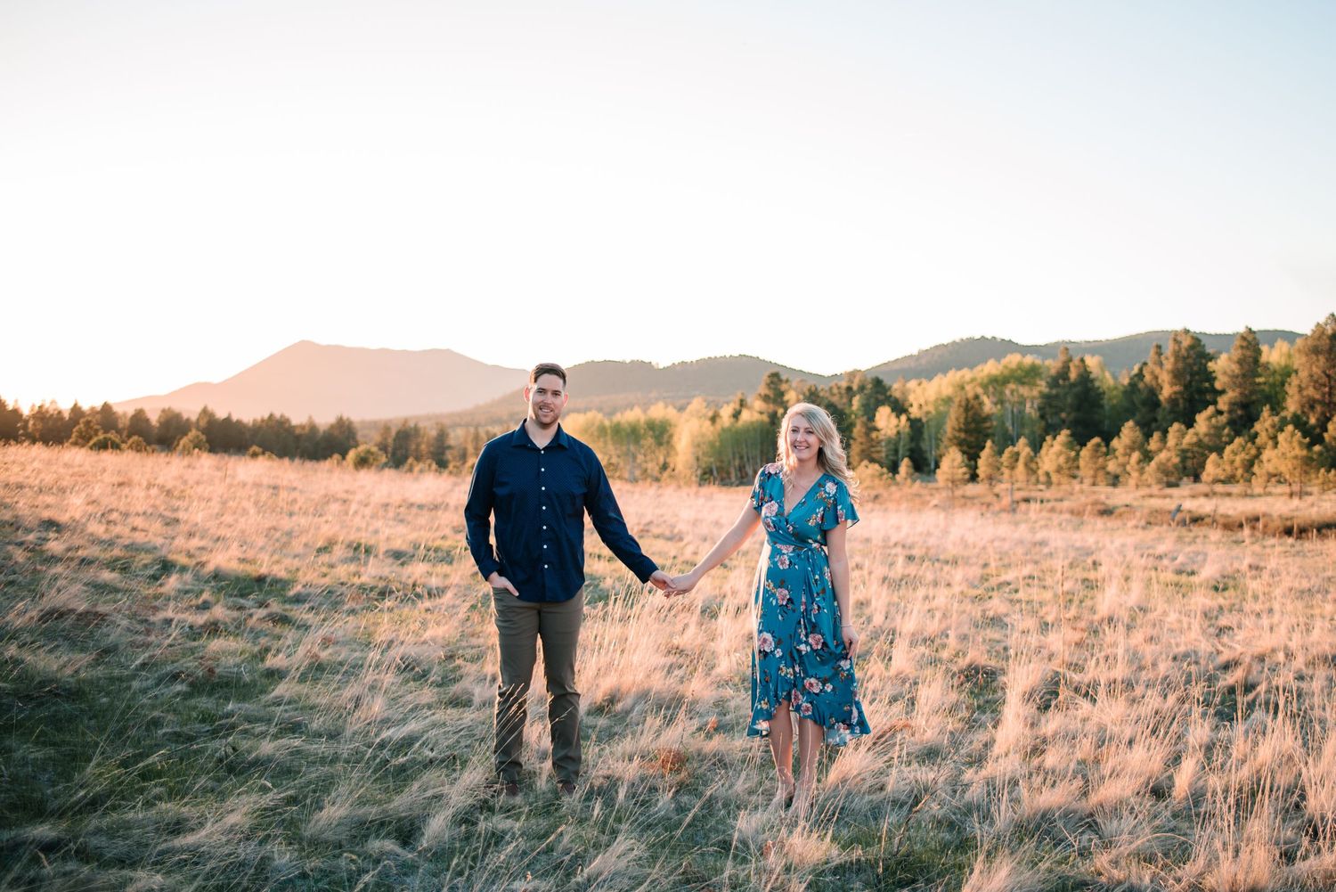 Couple walking hand in hand through tall grass field at sunset with mountains in background.
