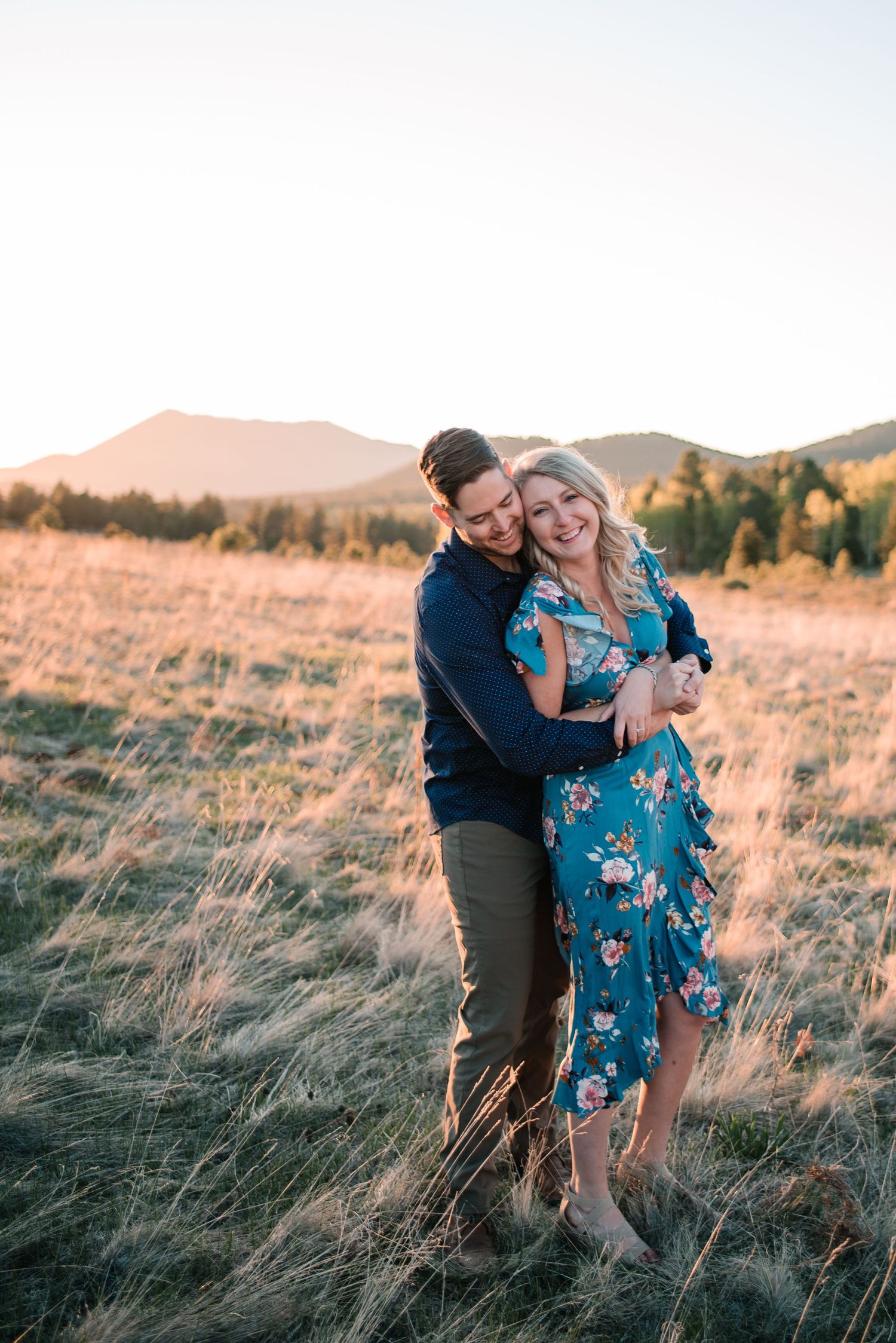 Couple embracing in golden field with mountains in background during sunset.