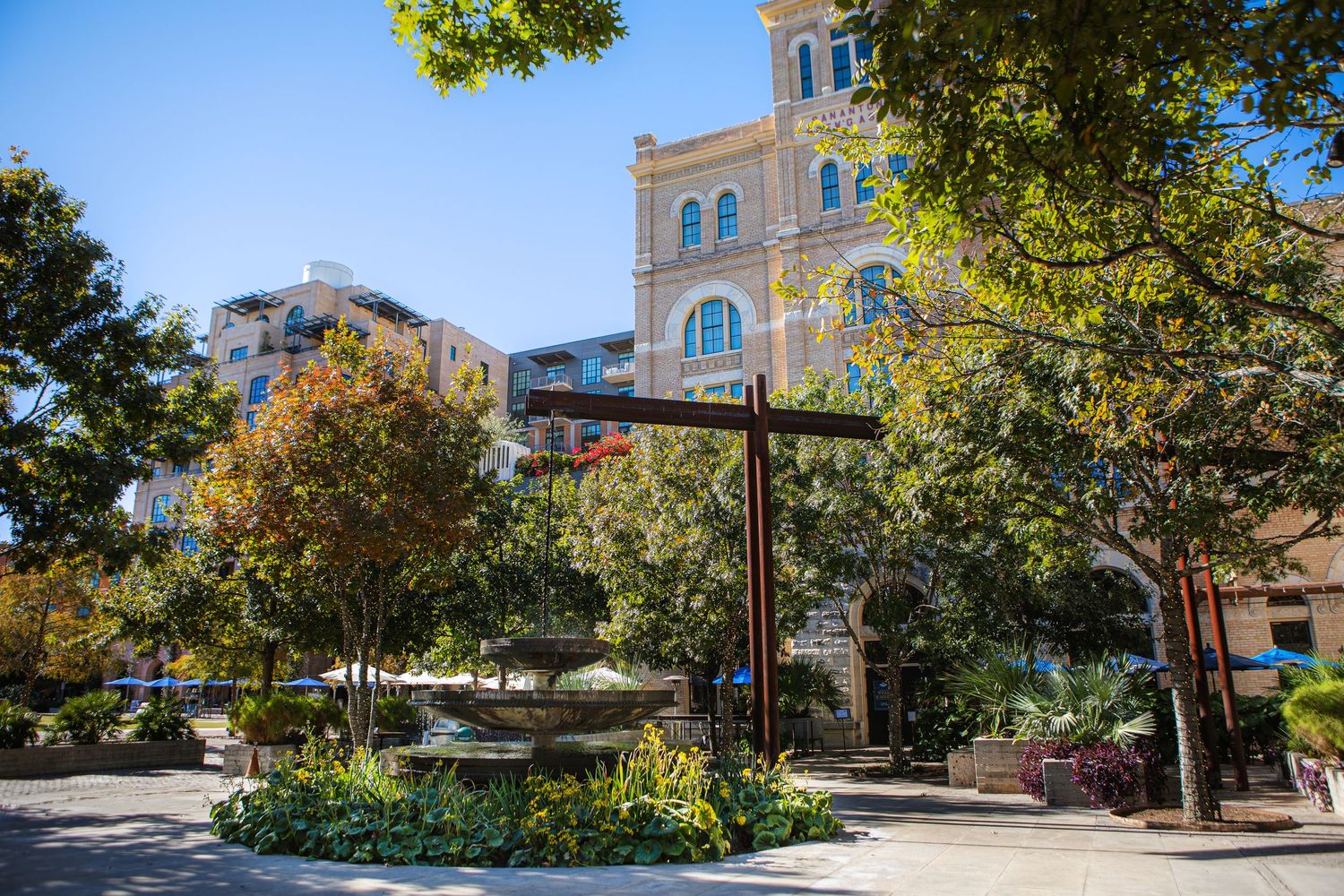 A large wooden cross stands in a garden area near a historic stone building on a sunny day.
