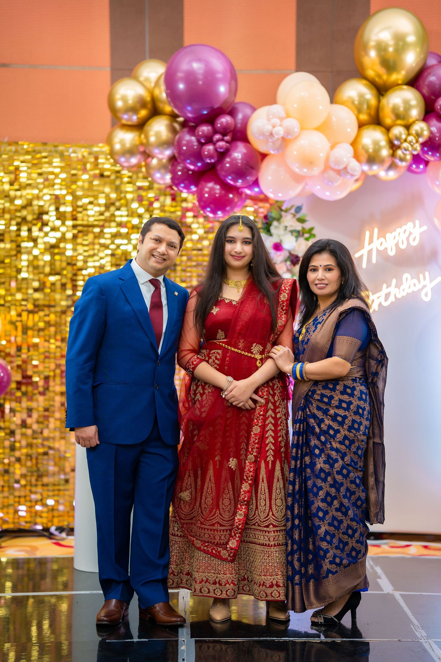 Family celebrating at a party with gold sequin backdrop and colorful balloon arch decorations.
