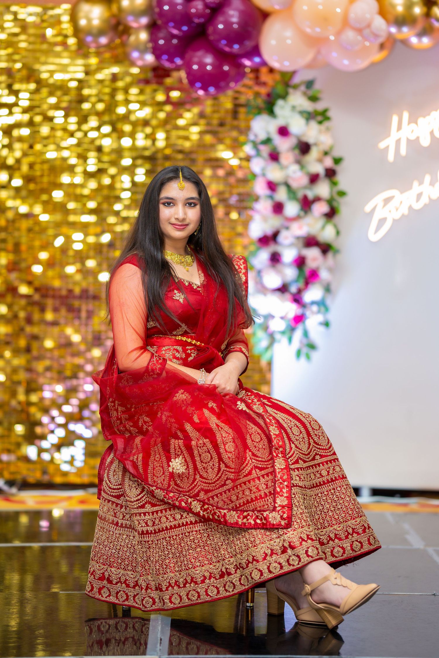 Elegant pose in traditional red lehenga against gold sequin wall with balloon decorations.