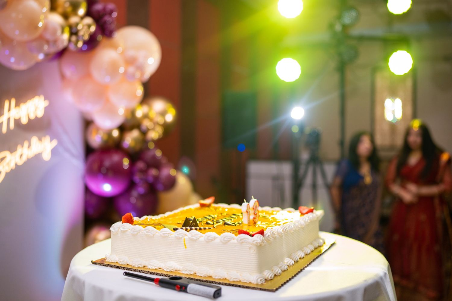 A white square wedding cake with floral decorations displayed at a reception with colorful lighting.