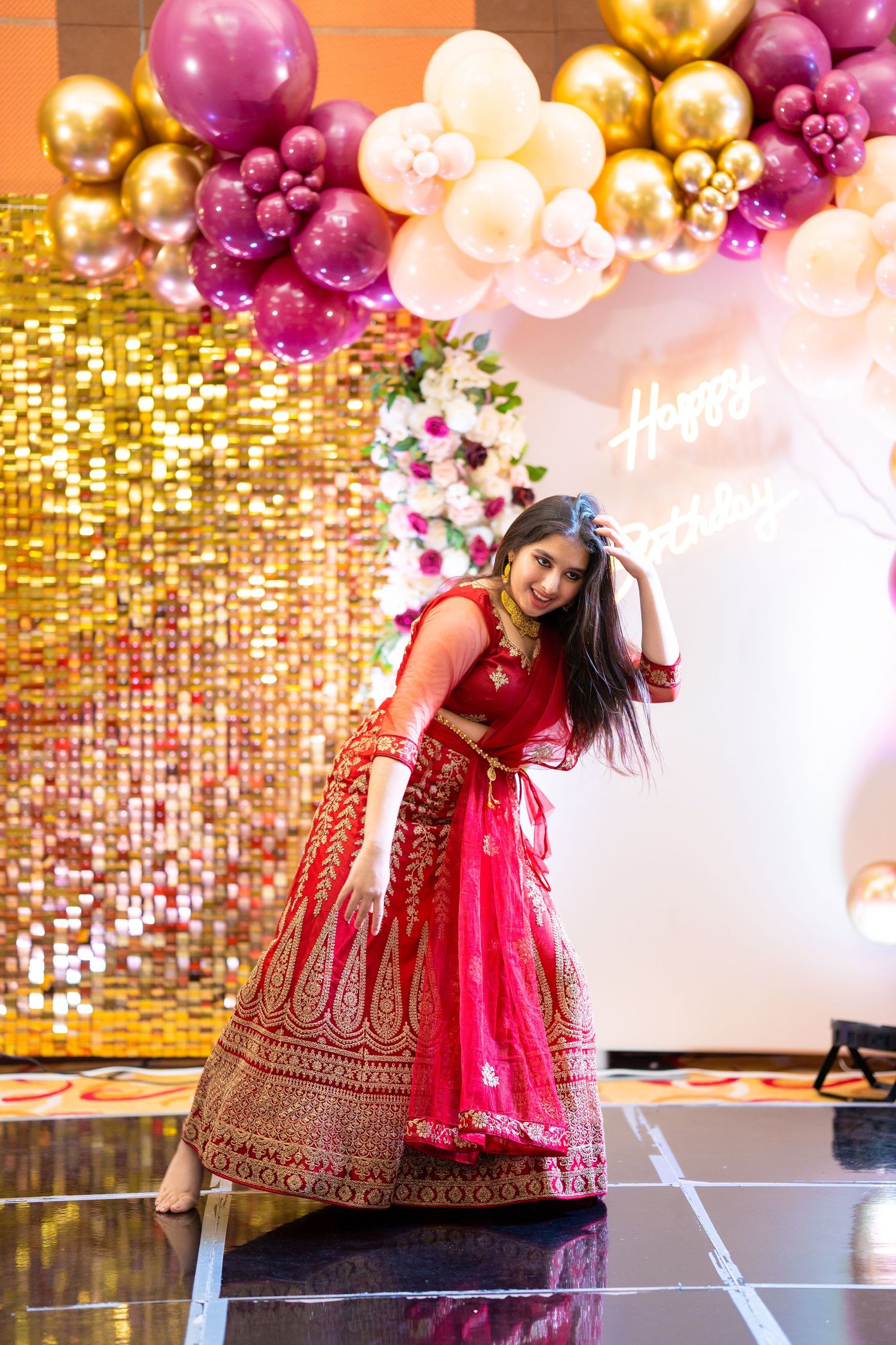 Celebratory dance pose in red lehenga against sparkling backdrop with balloon arch.