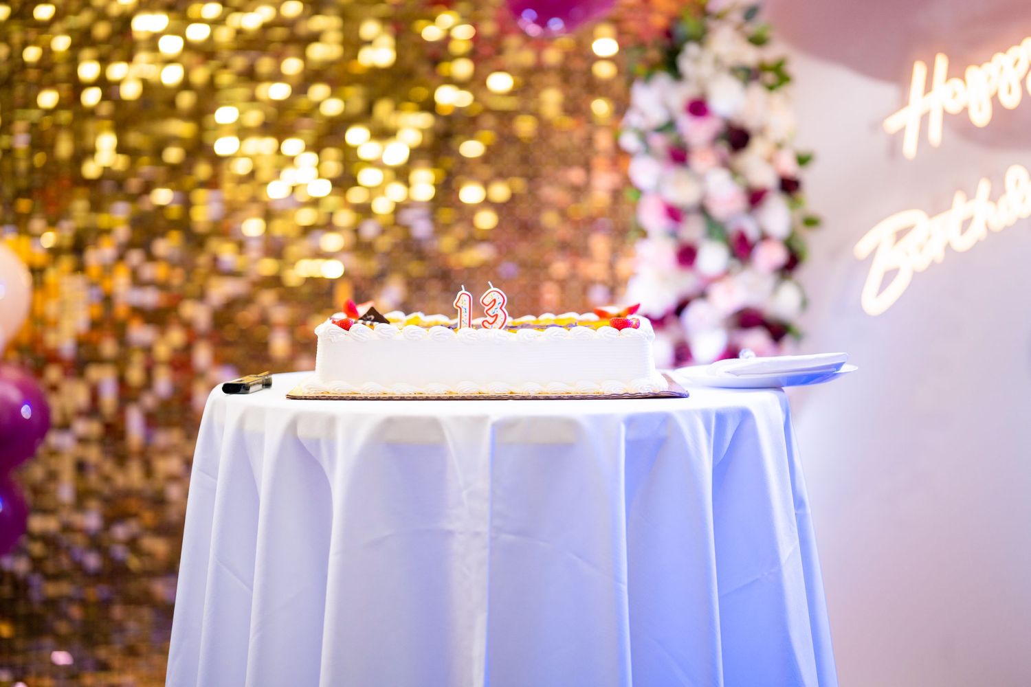 White tablecloth covered table with cake against gold sequin wall and balloon decorations.