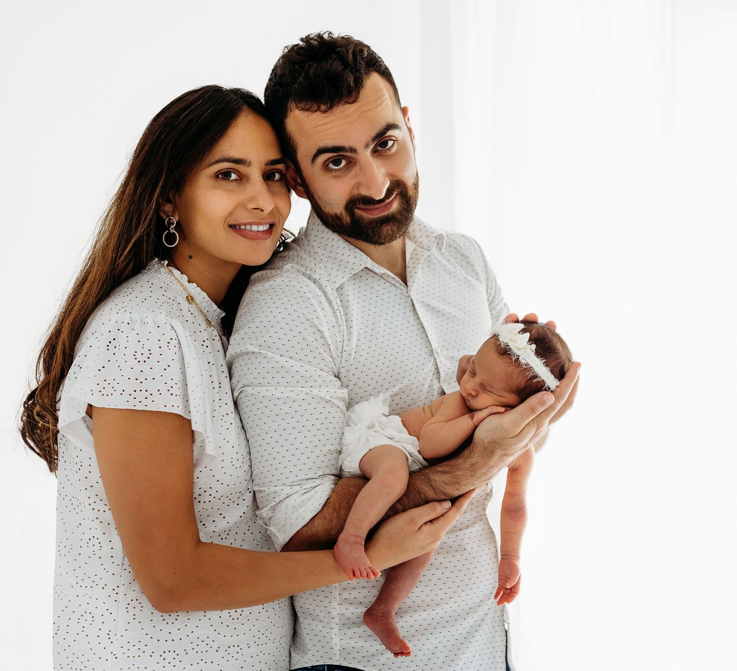 A family portrait featuring parents in matching white outfits holding their newborn baby.
