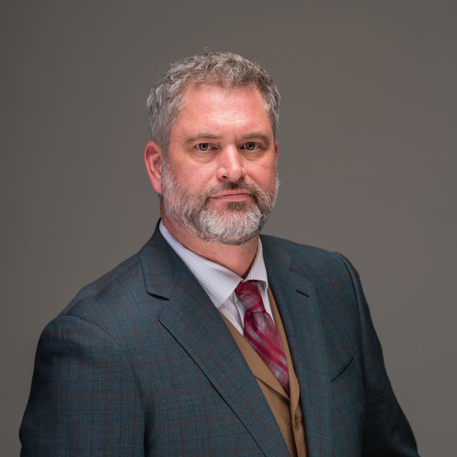 Professional headshot of person in navy suit with red tie and tan vest against gray background.