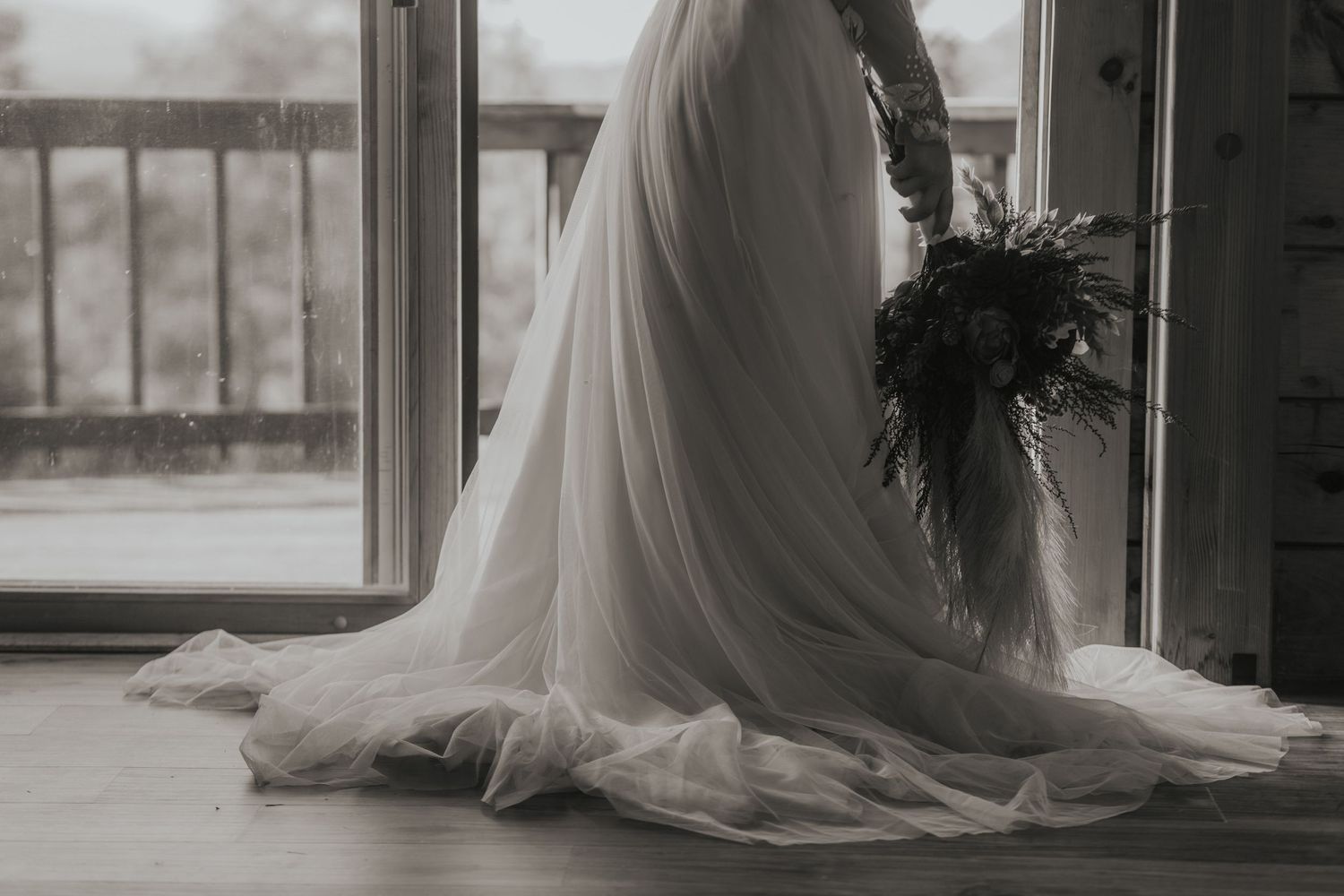 A bride in a flowing white gown stands silhouetted against a window in a moody black and white photographin New Hampshire NH USA