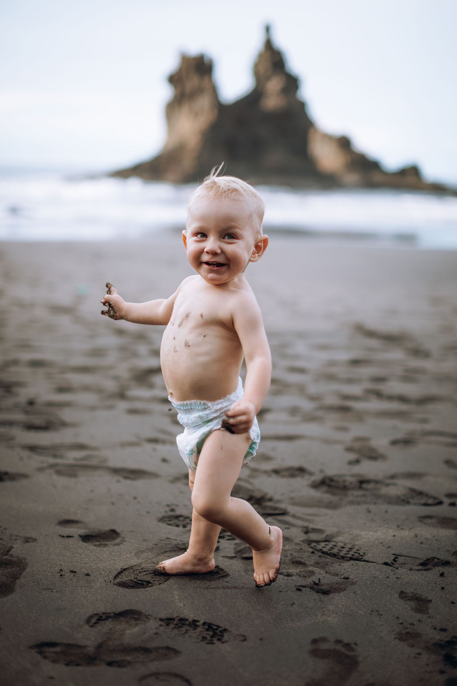 Toddler boy walking happily on the beach in his diaper with sand on his fingers and looking at the camera.
