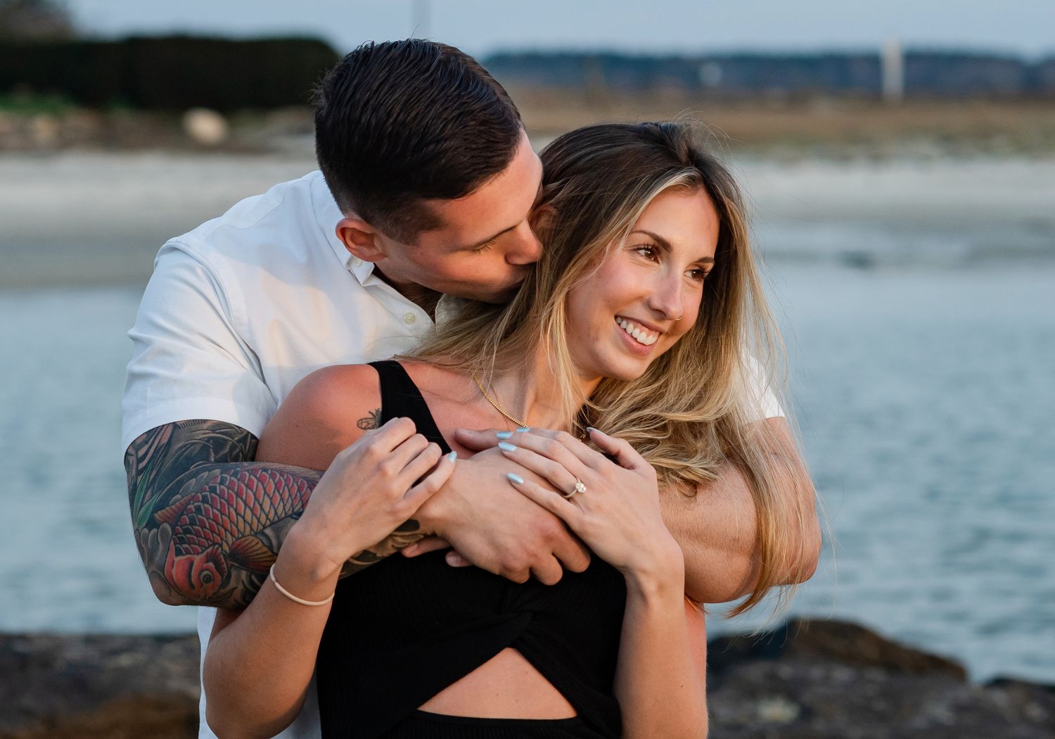 A couple embraces on a rocky beach during sunset with water in the background.