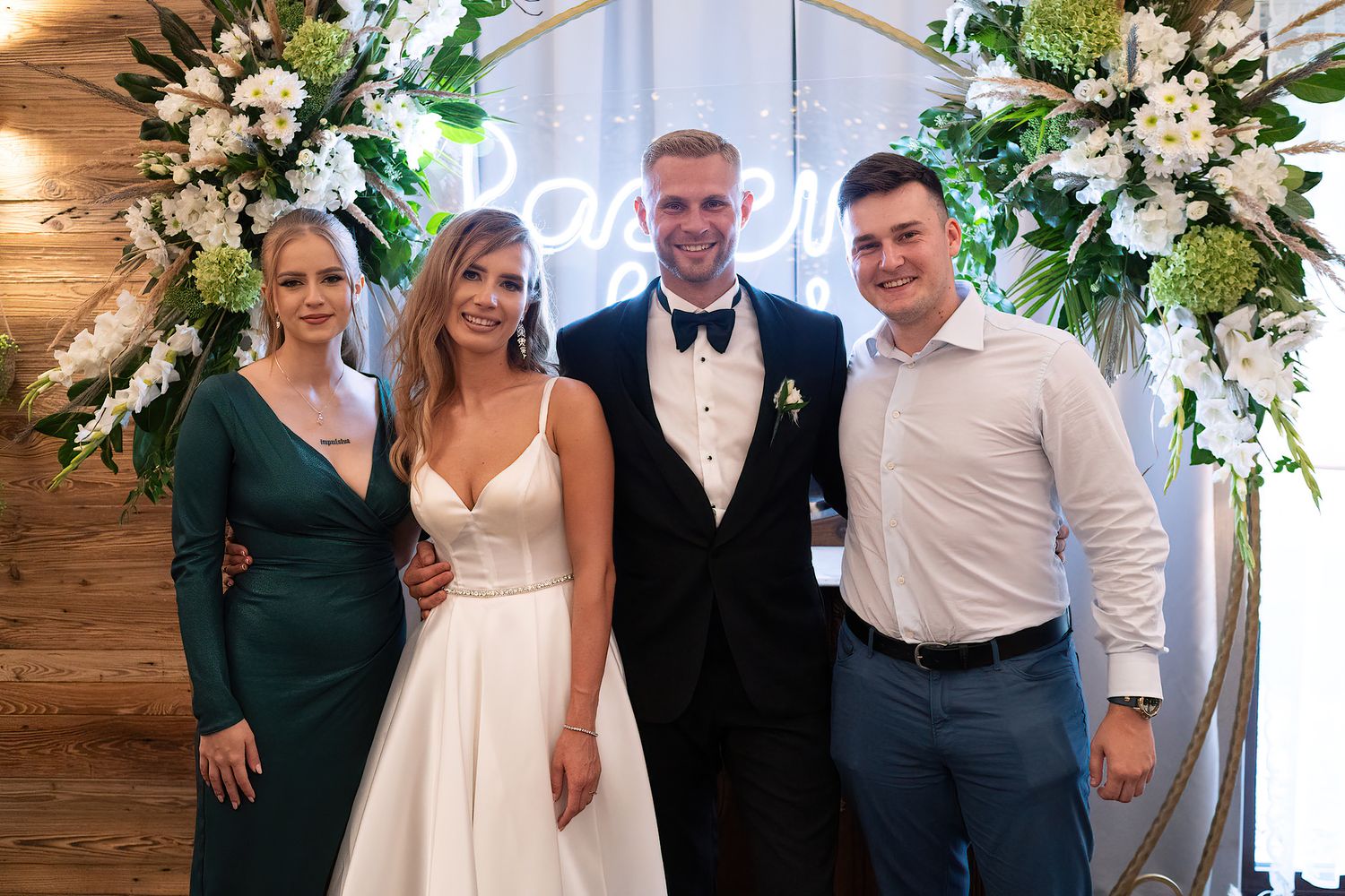 Wedding party poses under floral arch with white roses and greenery at indoor venue.