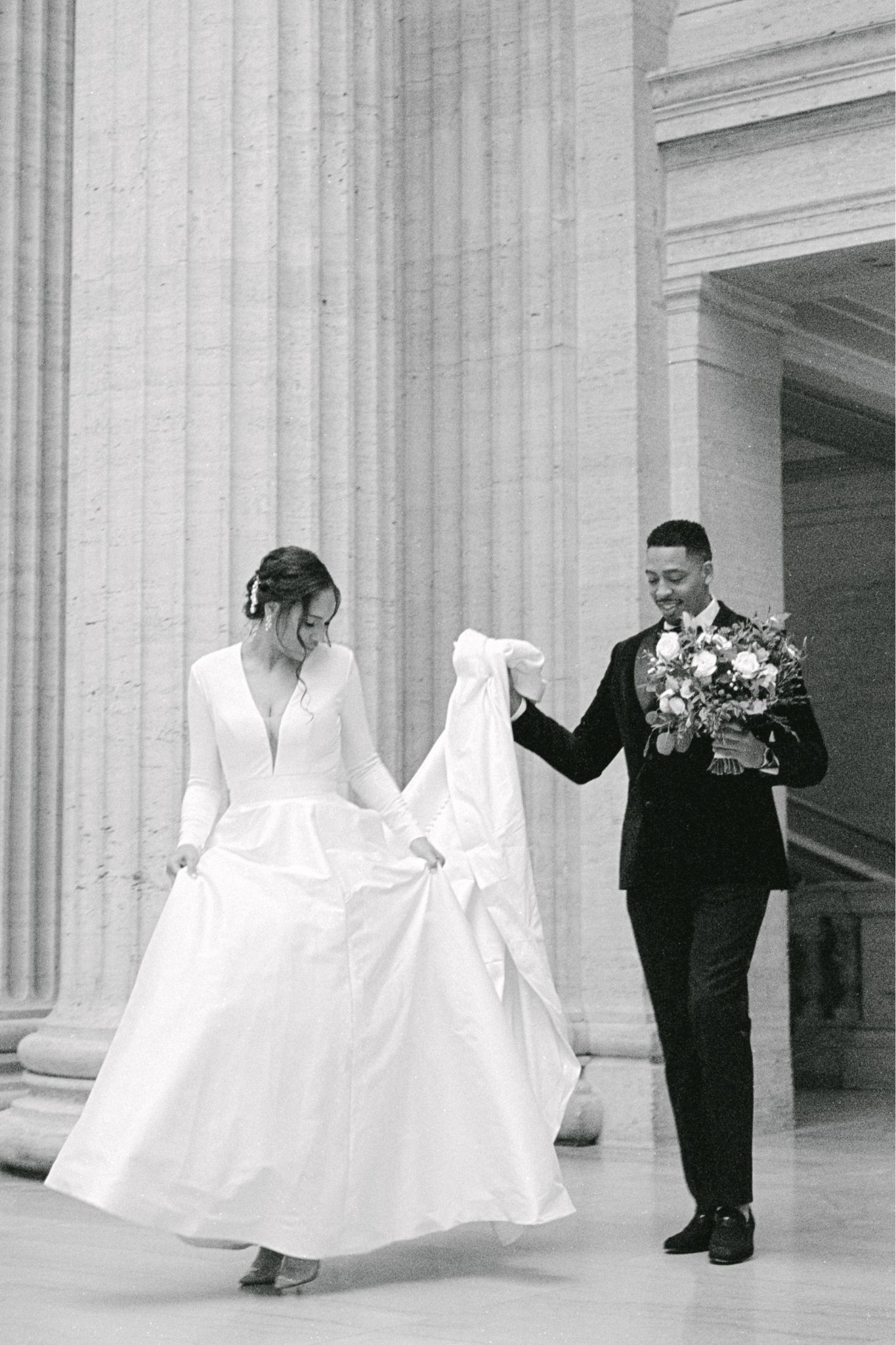 A couple in wedding attire dancing together outside a classical columned building.