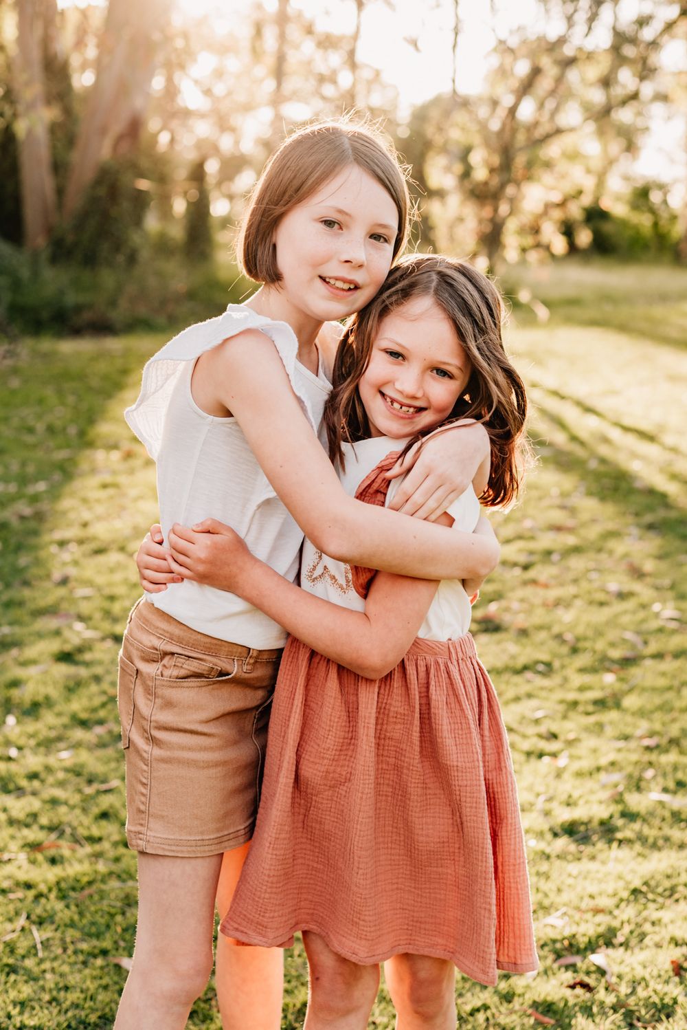 Two children wearing summer outfits embrace and smile while standing in golden evening sunlight.