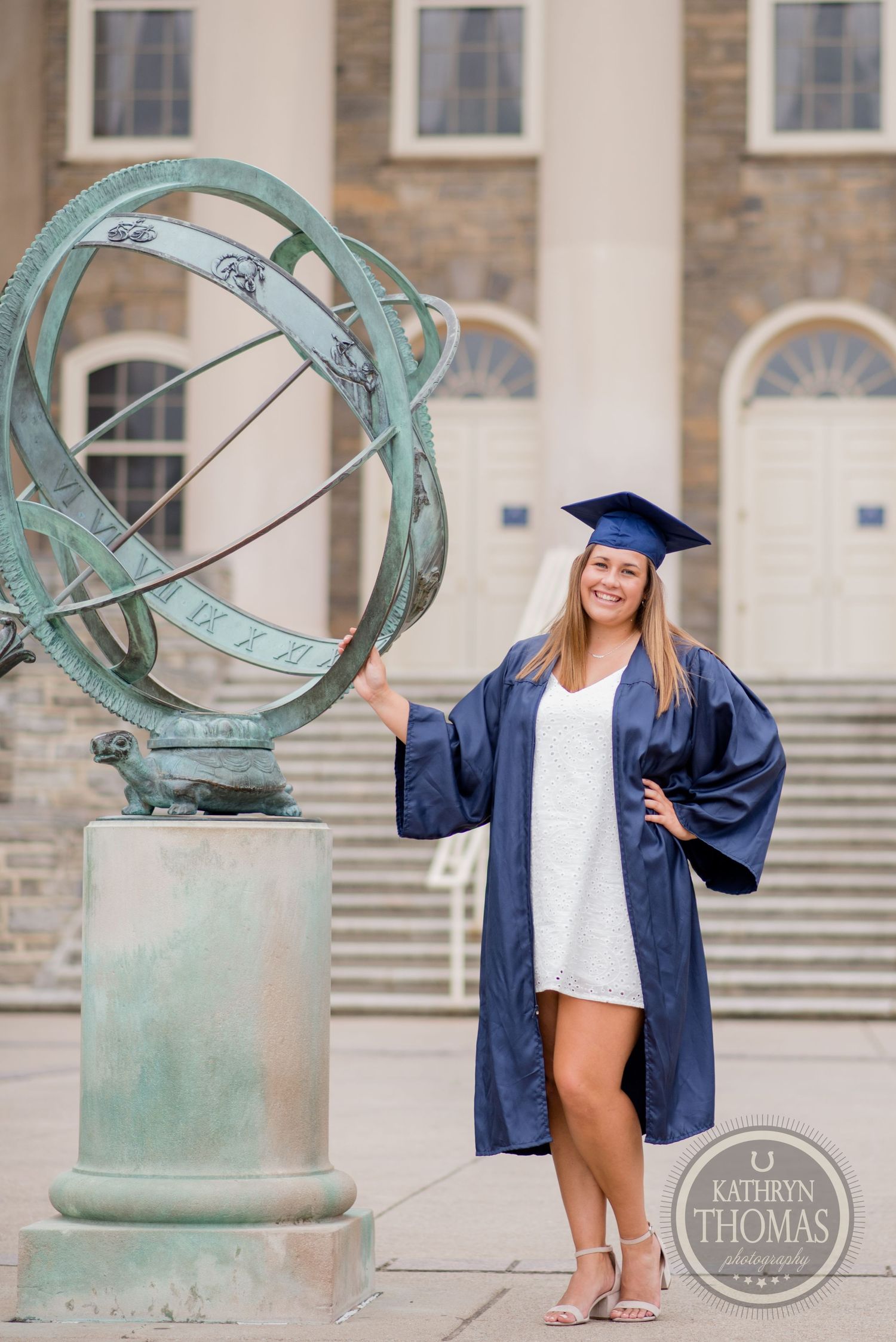 A graduate poses next to an ornate sundial sculpture outside a historic university building.