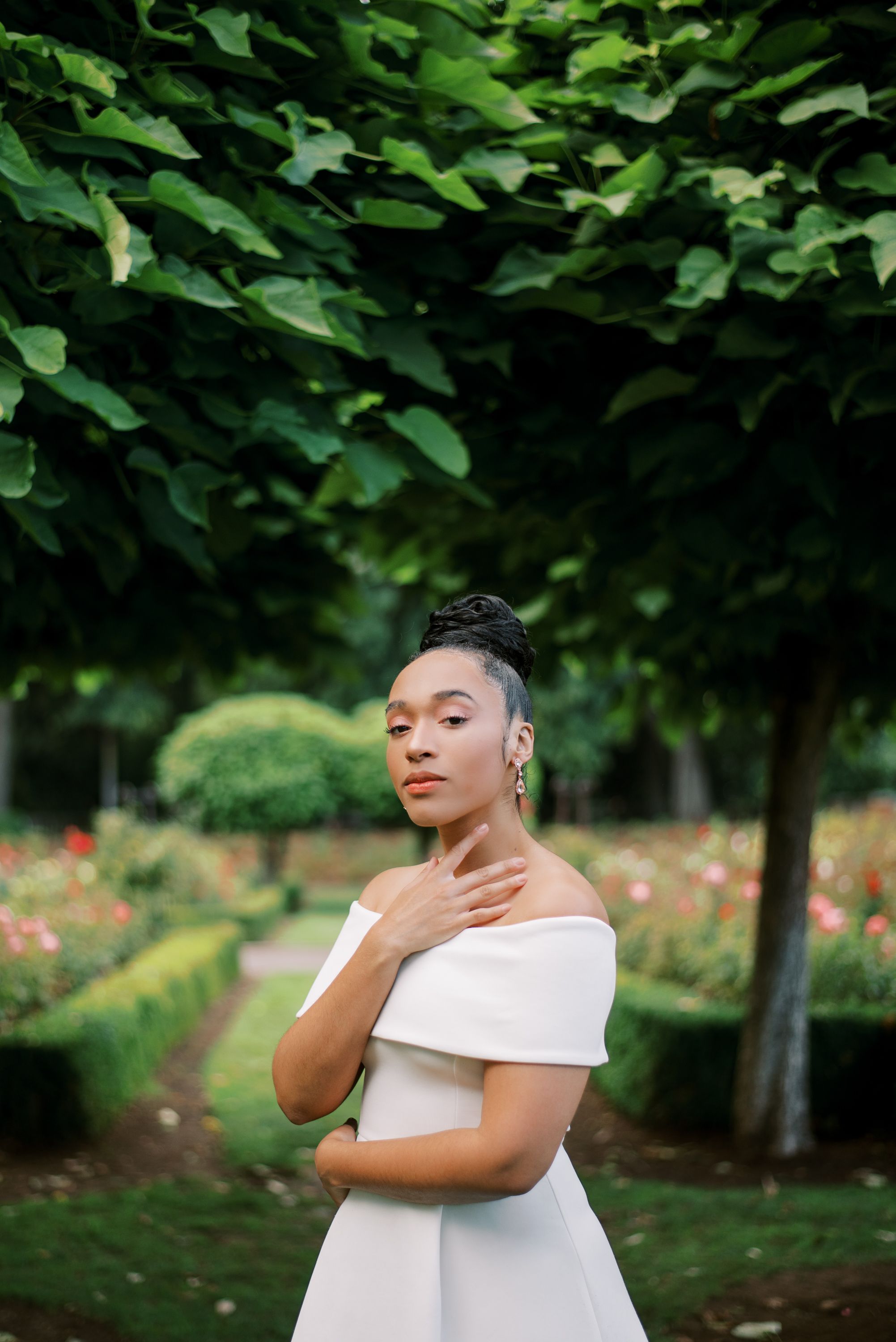 Elegant white off-shoulder dress photographed in a lush garden setting with roses and green trees.