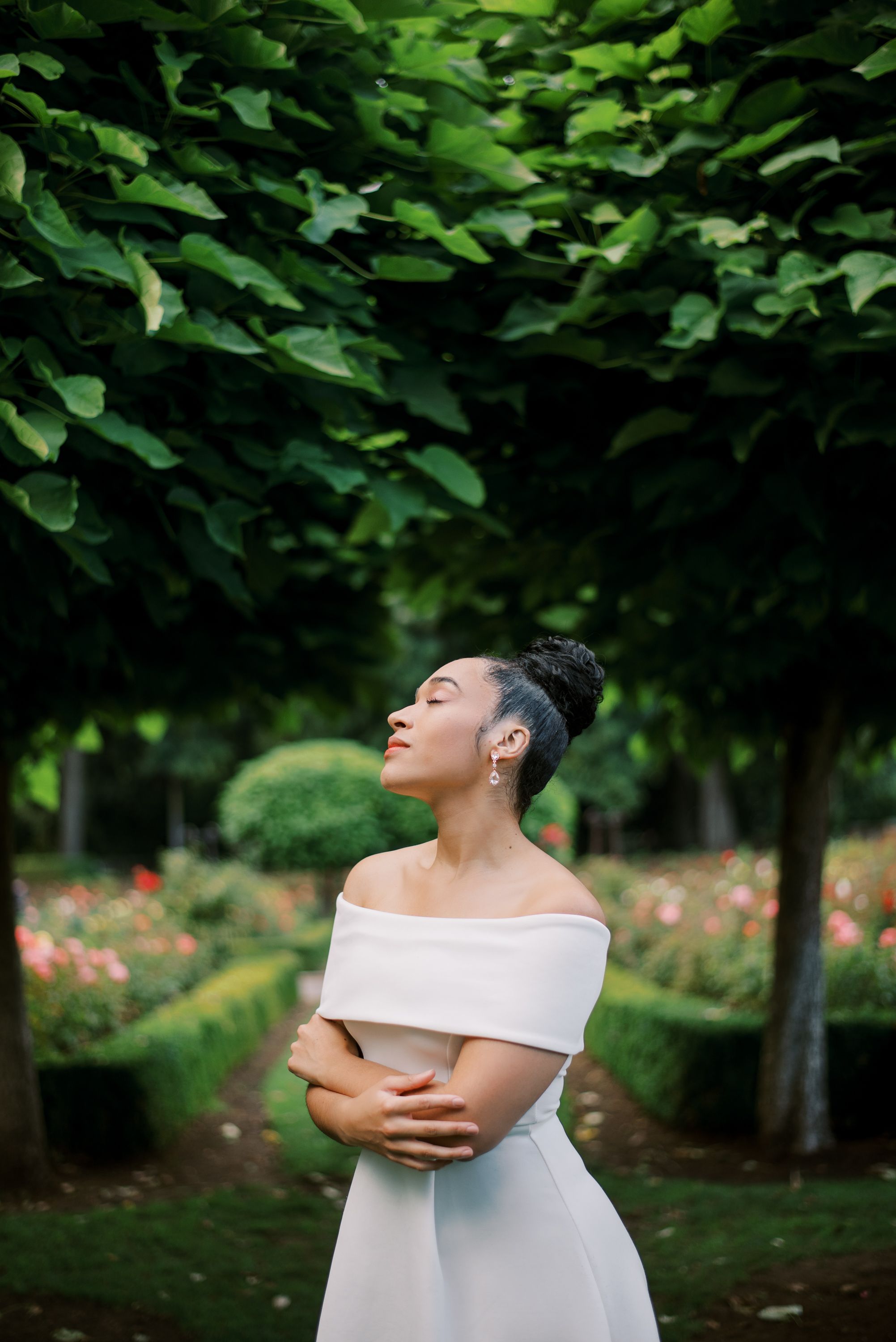 Off-shoulder white dress glows against dark green foliage in a romantic garden portrait.