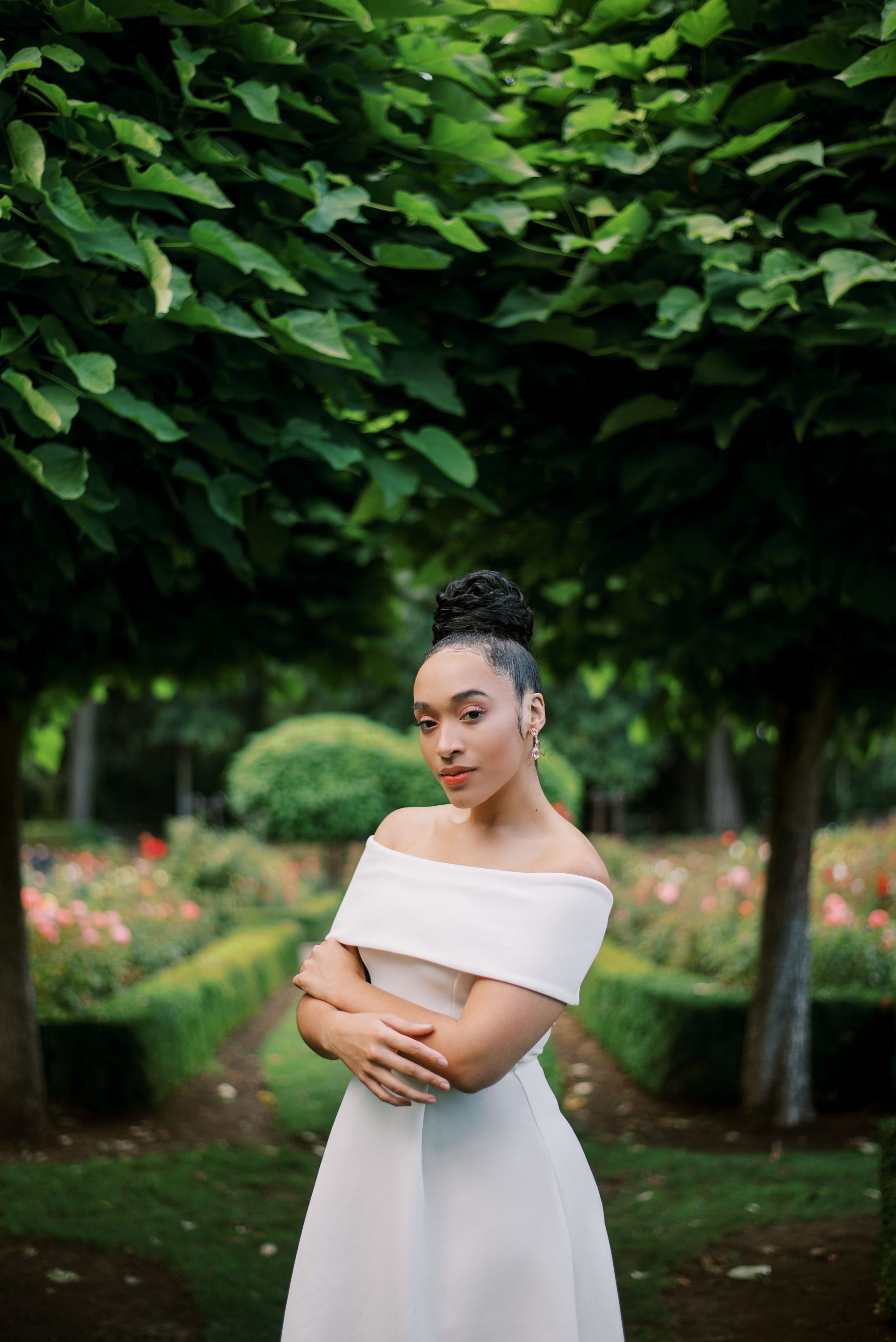 Classic white dress poses in a garden setting framed by green leaves and pink flowers.