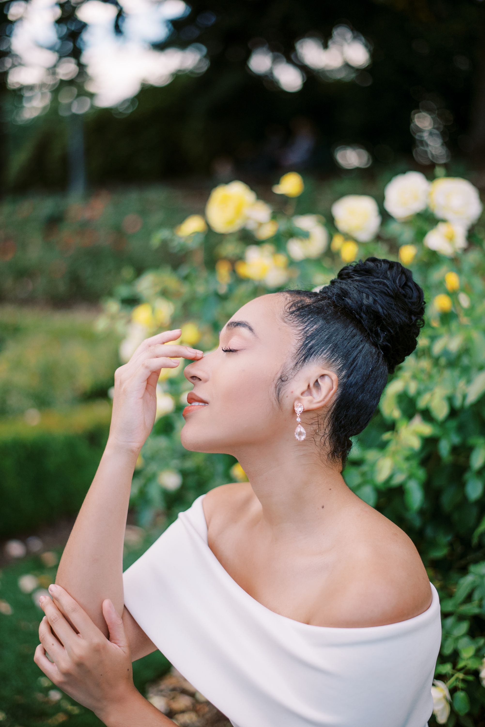 Graceful pose in white off-shoulder dress against yellow roses in a garden setting.