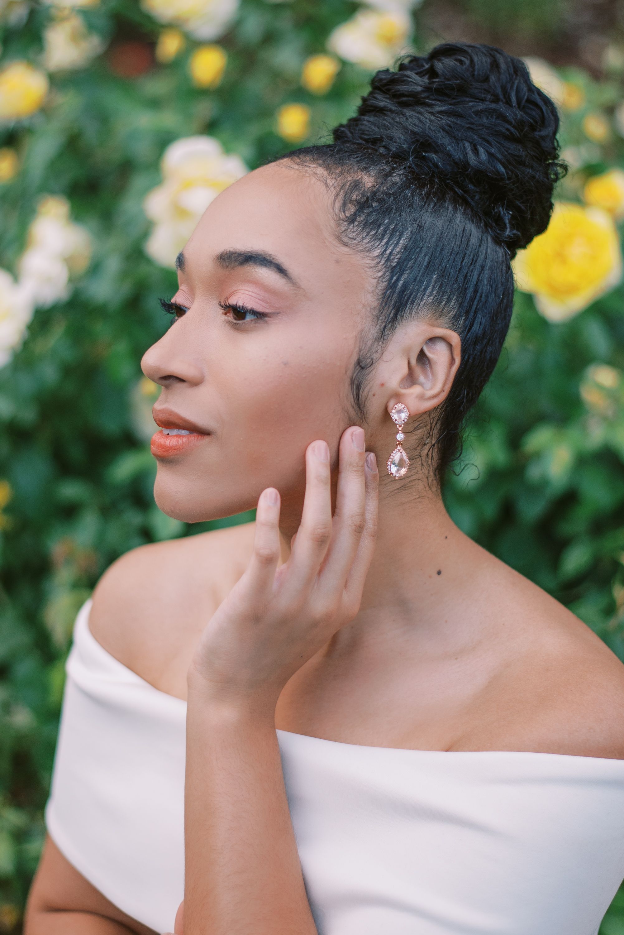 Elegant updo and crystal earrings complement a white off-shoulder dress against blooming flowers.