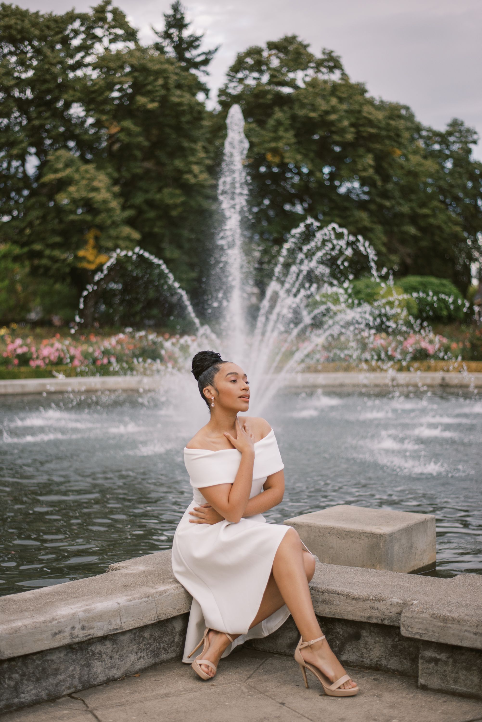 Elegant white dress portrait by fountain with water sprays and flowers in garden setting.