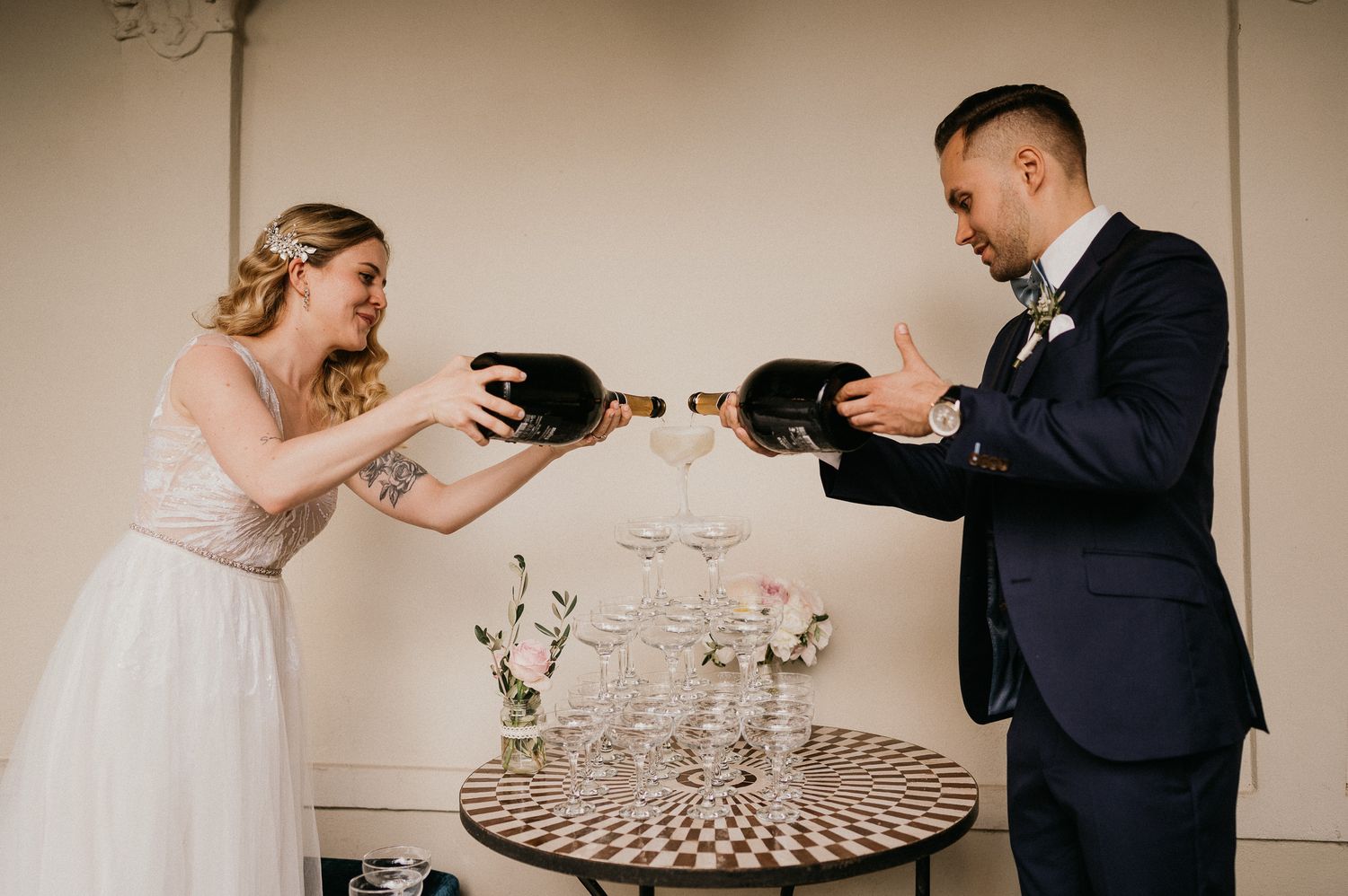 A playful wedding couple toasts with champagne bottles against a neutral wall.